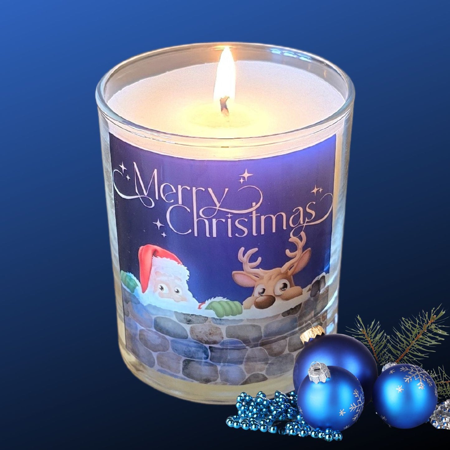 A lit Christmas scented candles with a charming Merry Christmas design featuring Father Christmas and a reindeer peeping over a wall. The candle is next to blue festive baubles.