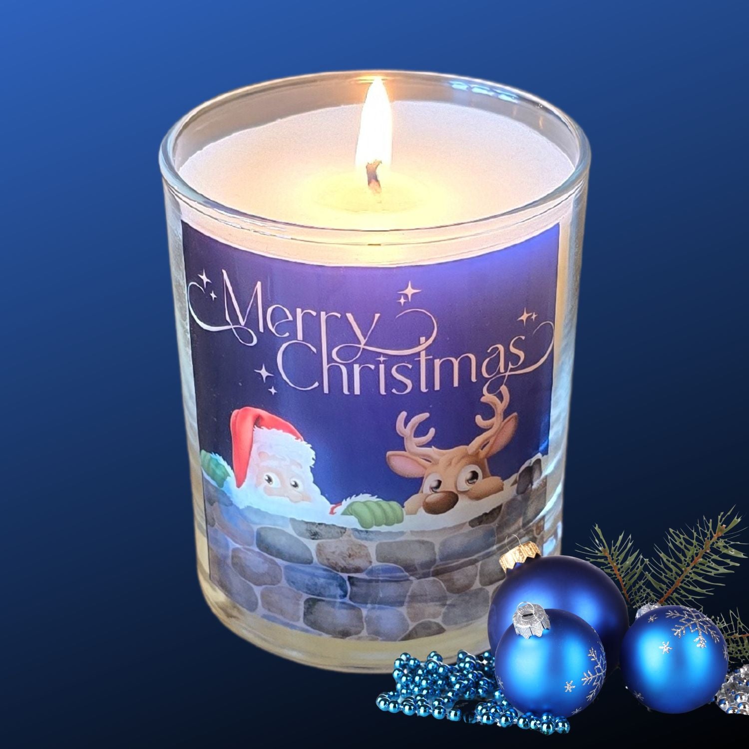 A lit Christmas scented candles with a charming Merry Christmas design featuring Father Christmas and a reindeer peeping over a wall. The candle is next to blue festive baubles.