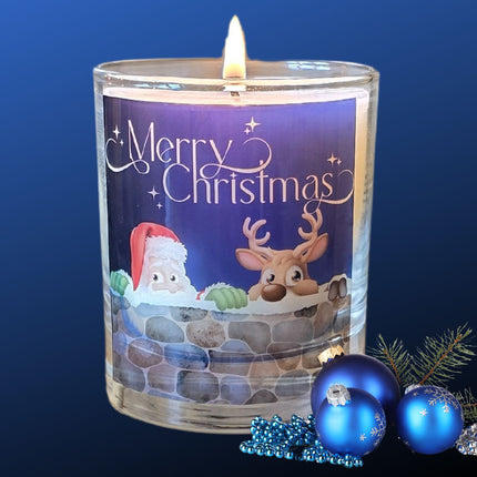A lit Christmas scented candles with a charming Merry Christmas design featuring Father Christmas and a reindeer peeping over a wall. The candle is next to blue festive baubles.