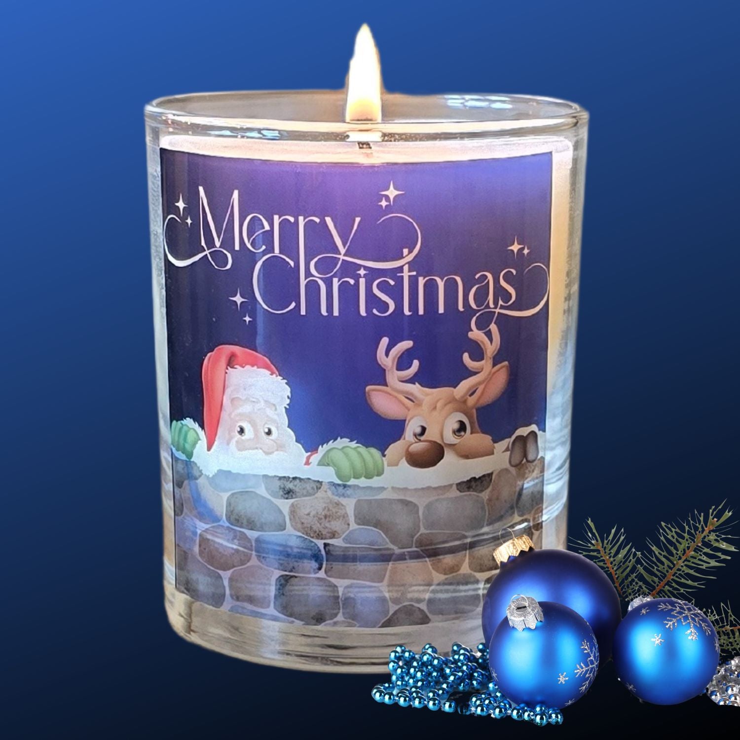 A lit Christmas scented candles with a charming Merry Christmas design featuring Father Christmas and a reindeer peeping over a wall. The candle is next to blue festive baubles.