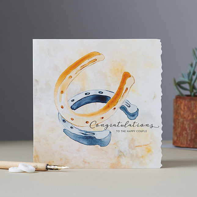 The Deckled Edge Ltd Equestrian Greeting Card Deckled Edge Wedding Or Anniversary features two watercolor horseshoes and “Congratulations to the happy couple” on the front, showcasing Janna Cossettini’s art.