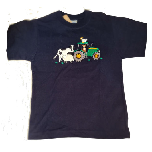 The British Country Collection Embroidered T Shirt features a black children’s tee with a colorful farmyard scene, showing a dog driving a green tractor and a white cow, sheep, and chicken nearby.