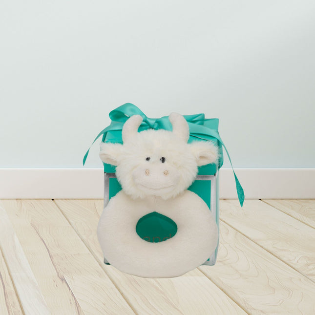 a donut shaped baby rattle in cream soft plush cream fabric with a cute cream highland cows head in front of a jomanda teal gift box in a room setting