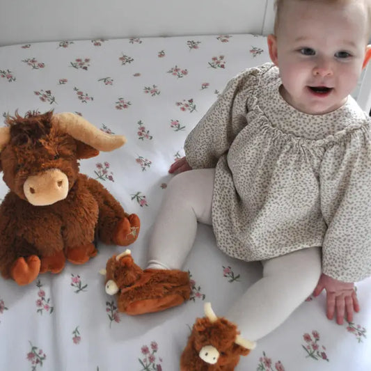 a baby with a highland cow soft toy and wearing a pair of fluffy brown baby boots featuring cute highland cow heads with cream nose and horns
