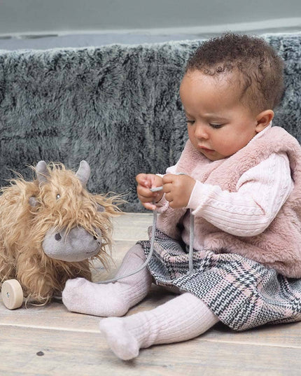 a cute highland cow stuffed toy with golden fur on wheels bing played with by a baby
