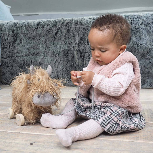 a cute highland cow stuffed toy with golden fur on wheels bing played with by a baby