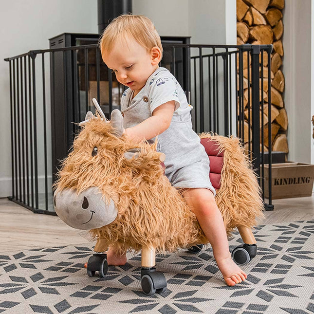 a baby riding a gorgeous highland cow ride on toy with golden fur, a loveable smiley face, leatherette seat and multi directional castors