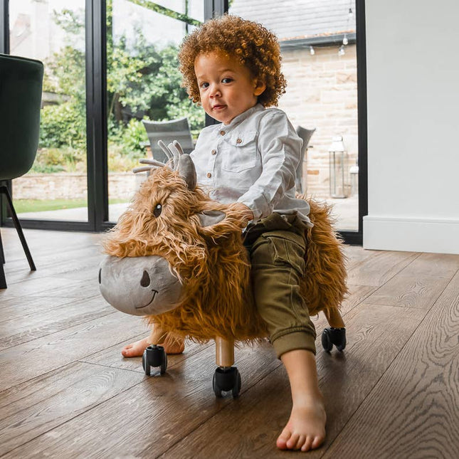 a toddler riding a gorgeous highland cow ride on toy with golden fur, a loveable smiley face, leatherette seat and multi directional castors
