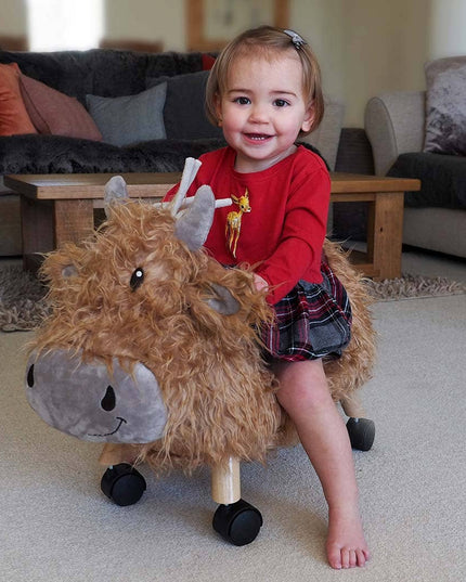 a baby girl riding a gorgeous highland cow ride on toy with golden fur, a loveable smiley face, leatherette seat and multi directional castors