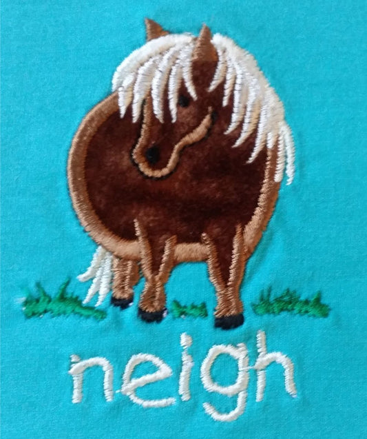 The British Country Collection Infant T Shirt features a cute embroidered pony with a white mane on green grass, neigh stitched below, and a blue 100% cotton background. Unisex design.