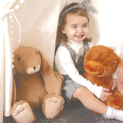 A smiling child in a plaid dress hugs the Jomanda Soft Toys Large Highland Cow Teddy Plush Baby Soft Toy beside a plush bunny inside a play tent; both toys are machine washable for easy care.