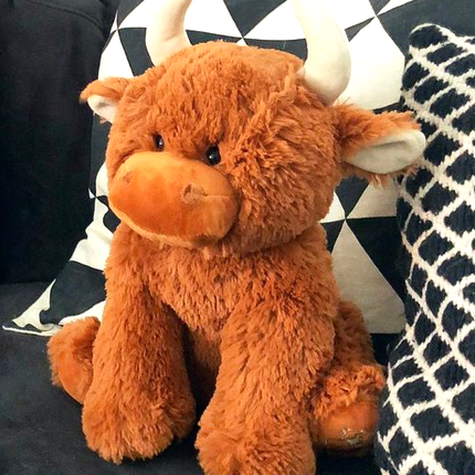 The Large Highland Cow Teddy Jomanda Plush Baby Soft Toy from Jomanda Soft Toys & Accessories sits on a dark couch with black-and-white pillows—an adorable, machine washable baby plush for easy care.