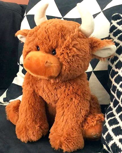 The Large Highland Cow Teddy Jomanda Plush Baby Soft Toy from Jomanda Soft Toys & Accessories sits on a dark couch with black-and-white pillows—an adorable, machine washable baby plush for easy care.