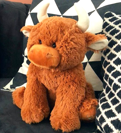 The Large Highland Cow Teddy Jomanda Plush Baby Soft Toy from Jomanda Soft Toys & Accessories sits on a dark couch with black-and-white pillows—an adorable, machine washable baby plush for easy care.