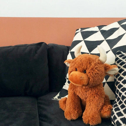 The Large Highland Cow Teddy Jomanda Plush Baby Soft Toy by Jomanda Soft Toys & Accessories, with white horns, rests on a dark sofa beside two patterned pillows. This adorable baby toy is machine washable. A salmon stripe decorates the wall behind.