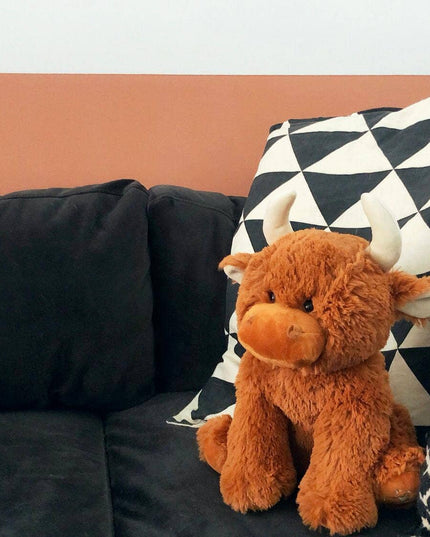 The Large Highland Cow Teddy Jomanda Plush Baby Soft Toy by Jomanda Soft Toys & Accessories, with white horns, rests on a dark sofa beside two patterned pillows. This adorable baby toy is machine washable. A salmon stripe decorates the wall behind.