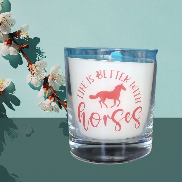 The Quaver&Lyric Scented Candle In Glass Container features a Life is Better With Horses design and horse image, presented on a reflective surface with white blossoms against a light blue backdrop and infused with premium fragrances.