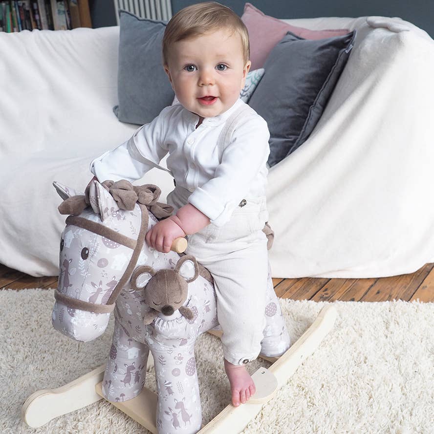 a happy baby riding a cute fabric rocking horse with brown bridle and mane on wooden rockers with a soft toy mouse in a pocket on the horses shoulder