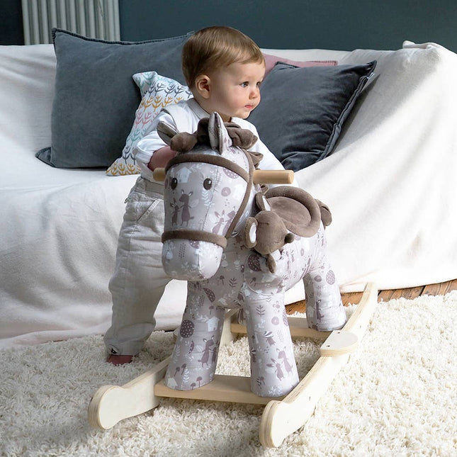 a baby standing next to a cute fabric horse with brown bridle, saddle, mane and tail on wooden rockers with a soft toy mouse in a pocket on its shoulder