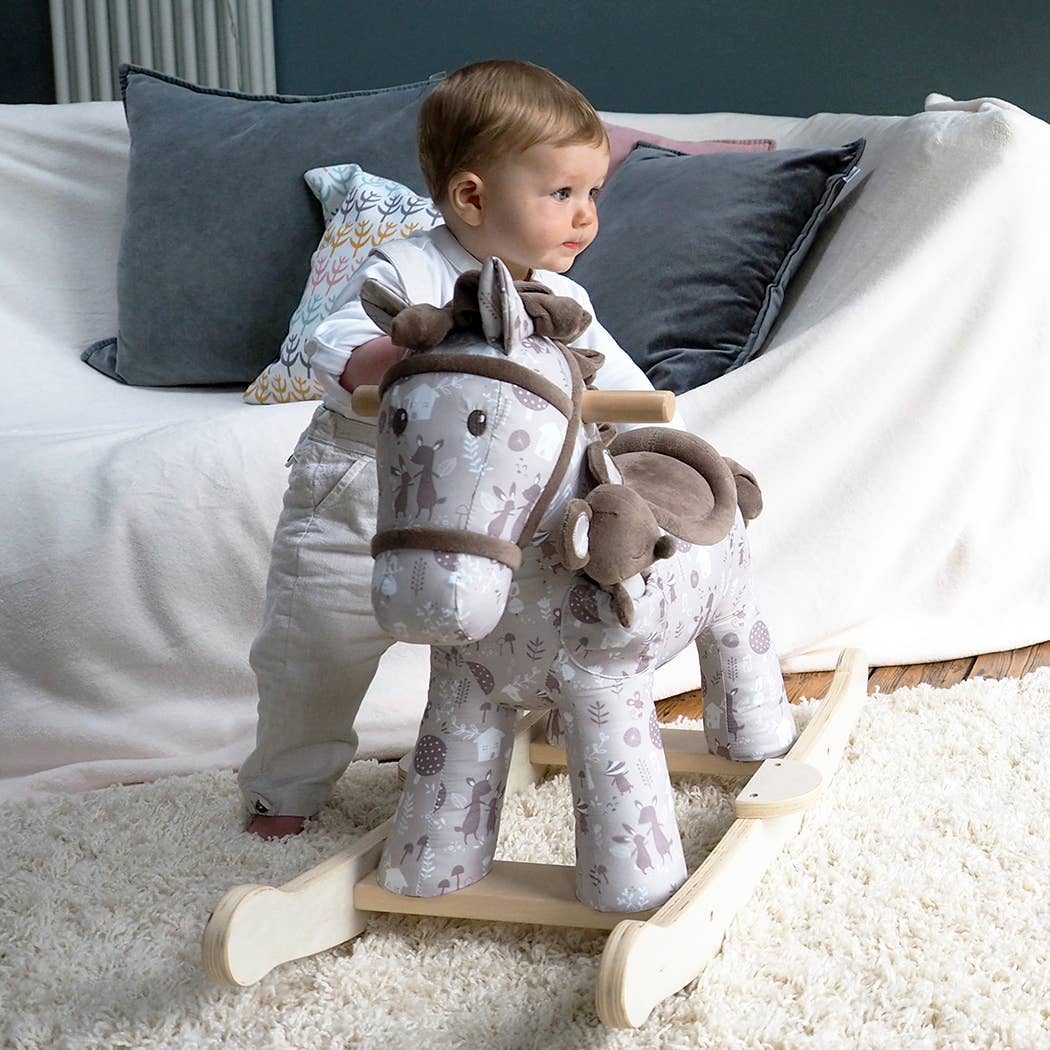 a baby standing next to a cute fabric horse with brown bridle, saddle, mane and tail on wooden rockers with a soft toy mouse in a pocket on its shoulder