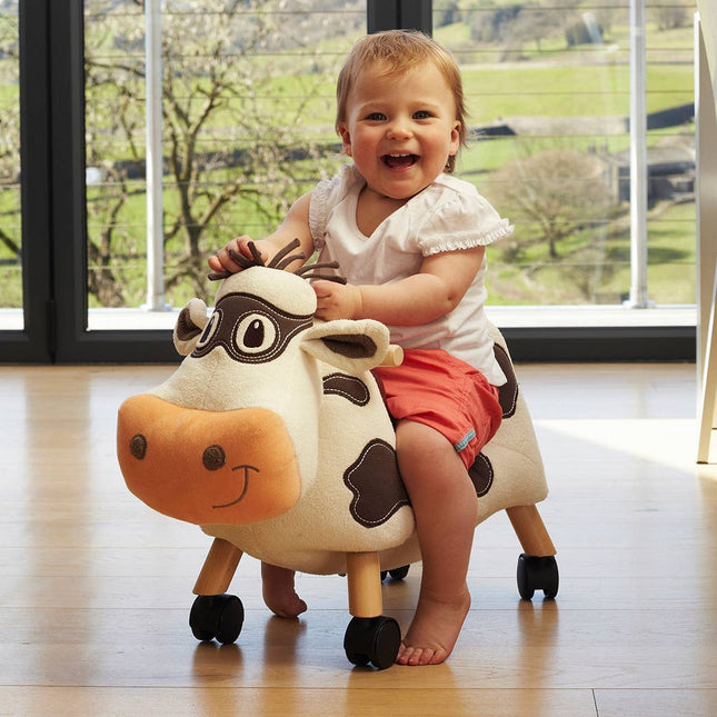 A smiling baby in a white shirt and pink shorts enjoys the Little Bird Told Me UK Moobert Cow Ride On Toy—a charming wooden toy with a padded cow-shaped seat, great for encouraging toddler development indoors by scenic windows.