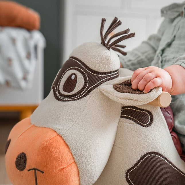 A child in light green holds the ear of the Moobert Cow Ride On Toy by Little Bird Told Me UK, a plush, cartoon-style wooden ride-on with a padded seat, soft fabric, large stitched eyes, and an orange snout.