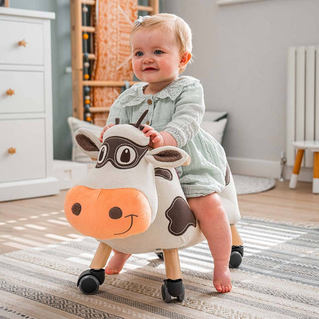 A smiling baby in a light green dress enjoys the Moobert Cow Ride On Toy by Little Bird Told Me UK, a whimsical wooden toy on wheels with a padded seat, perfect for encouraging toddler development in any cozy play space.