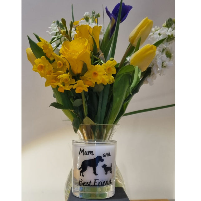 A Quaver&Lyric Scented Candle in a 20cl glass container features a Mum and Best Friend dog designâ€”an ideal gift for dog lovers. Displayed with yellow tulips, daffodils, and purple irises in a glass vase.