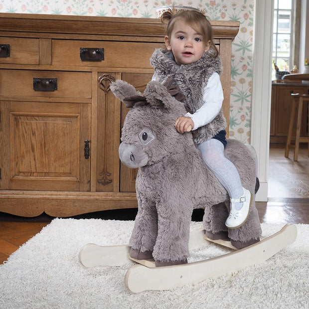 A young girl enjoys imaginative play on the Little Bird Told Me UK Norbert Rocking Donkey, surrounded by wooden furniture and floral wallpaper. She wears a textured taupe coat, white shirt, tights, and white shoes.