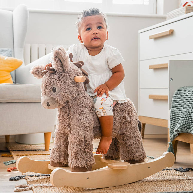 A toddler in a white shirt enjoys the Norbert Rocking Donkey by Little Bird Told Me UK—a plush wooden toy—while seated in a cozy, modern room designed for imaginative play.