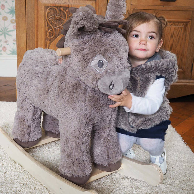 A young child hugs the Norbert Rocking Donkey from Little Bird Told Me UK in a cozy room with wooden furniture and a light rug—perfect for imaginative play with this charming wooden children’s toy.
