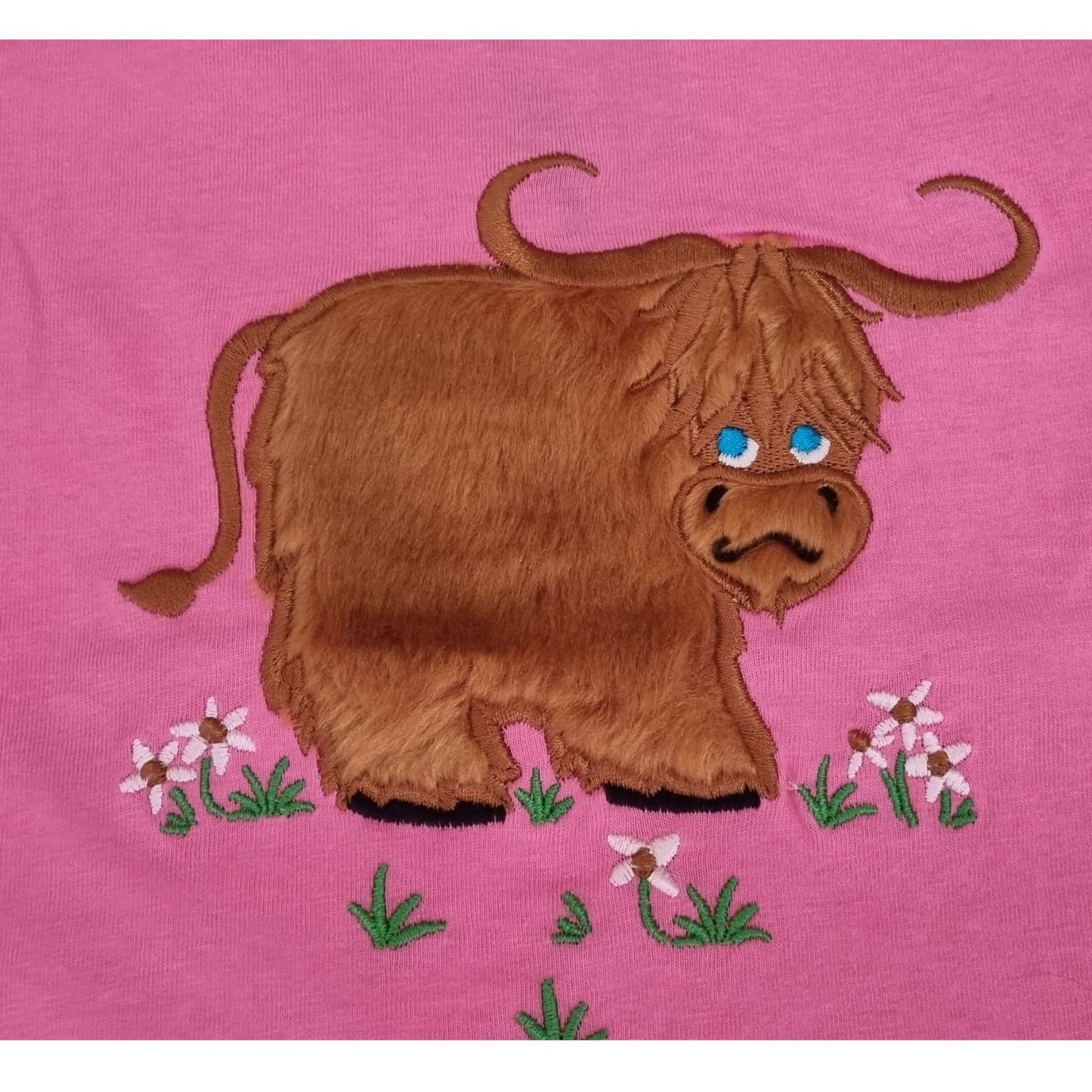 The Ramblers Highland Cow Embroidered T Shirt MacMoo Design features a shaggy brown cow with blue eyes on pink 100% cotton, standing on grass with pink and white flowersâ€”a cute childrens tee.