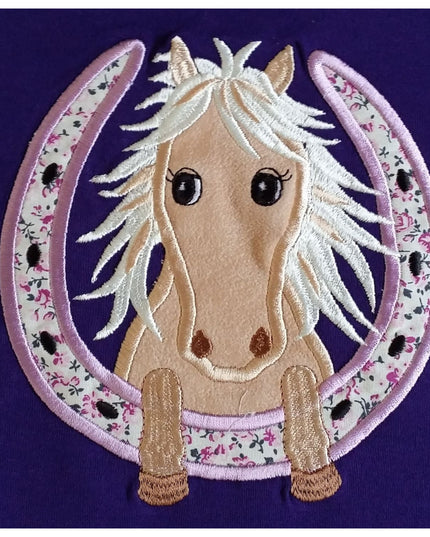 The British Country Collection Horse Embroidered T Shirt Childrens Purple Horseshoe features a light brown horse with a white mane and big black eyes inside an embroidered horseshoe, plus small flowers and tiny black horses on 100% cotton.