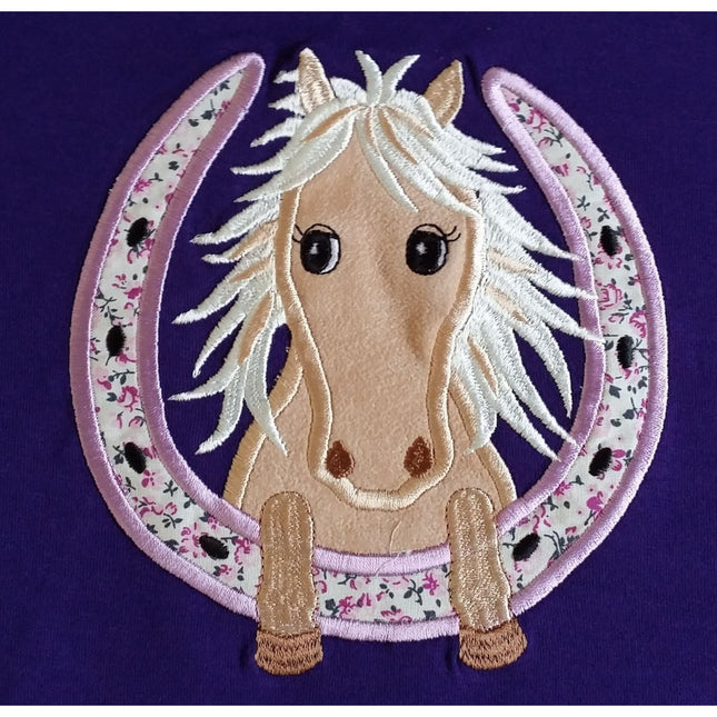 The British Country Collection Horse Embroidered T Shirt Childrens Purple Horseshoe features a light brown horse with a white mane and big black eyes inside an embroidered horseshoe, plus small flowers and tiny black horses on 100% cotton.