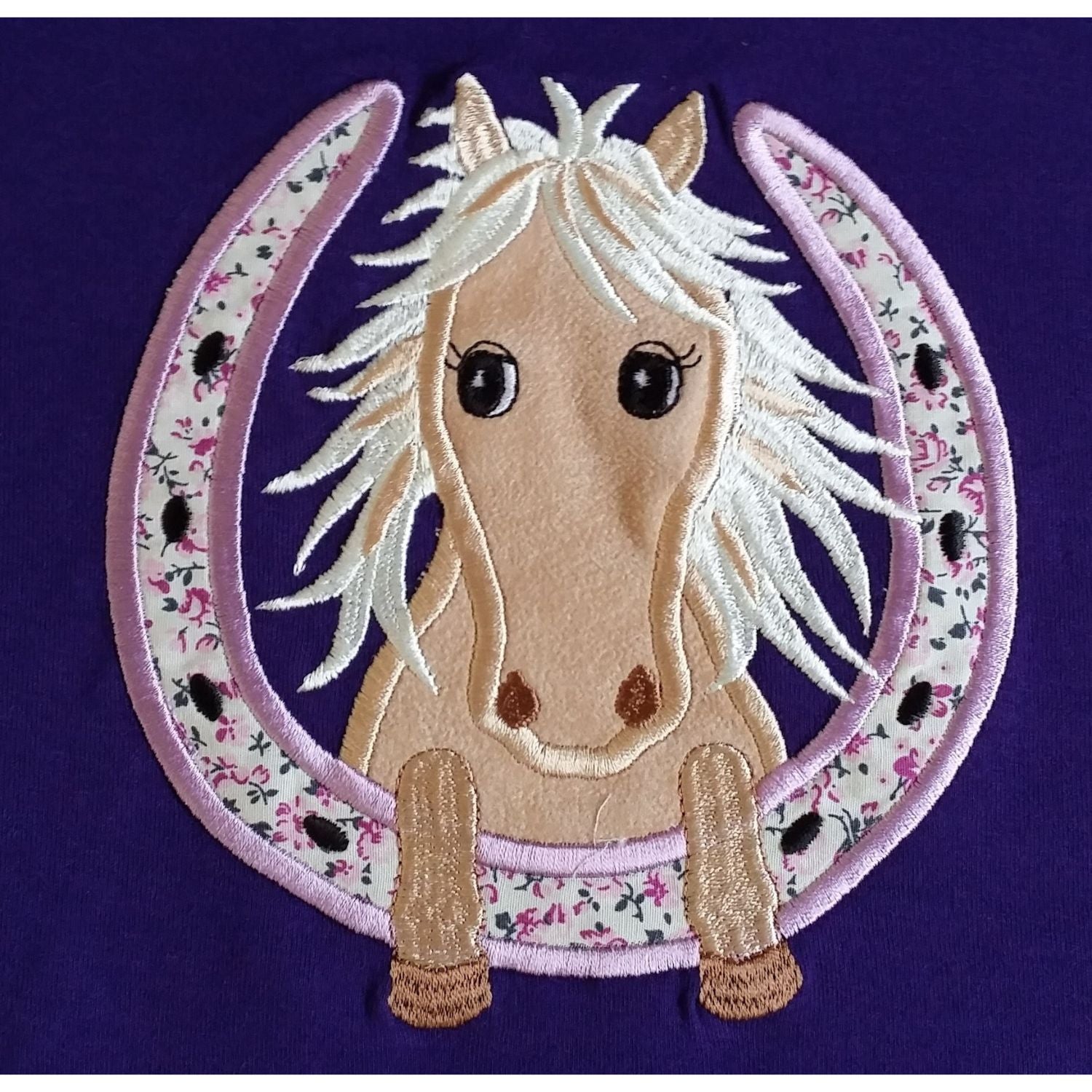 The British Country Collection Horse Embroidered T Shirt Childrens Purple Horseshoe features a light brown horse with a white mane and big black eyes inside an embroidered horseshoe, plus small flowers and tiny black horses on 100% cotton.