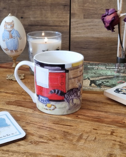 The Dog Mug Alisons Animals Pre-wash Cartoon Gift Boxed by Alisons Animals, with a fun cat and dog illustration, sits among books, dried roses, a candle, an egg, and cat-themed cards—an ideal gift for animal lovers.