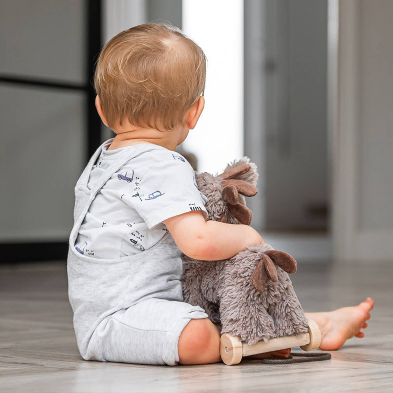 A toddler sits on a wooden floor, playing with the Pull Along Baby Toy Donkey Norbert by Little Bird Told Me UK. Facing away, the child wears a light romper and printed shirt, enjoying fun motor skills development.