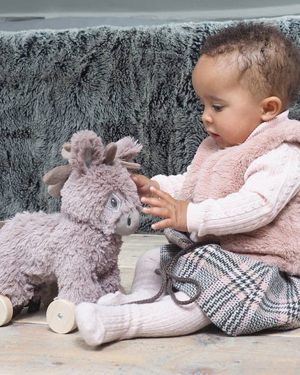 A baby in a pink vest, plaid skirt, and tights sits on a wooden floor, reaching for the Little Bird Told Me UK Norbert Pull Along Baby Toy Donkey to foster motor skills. A fuzzy gray blanket rests in the background.