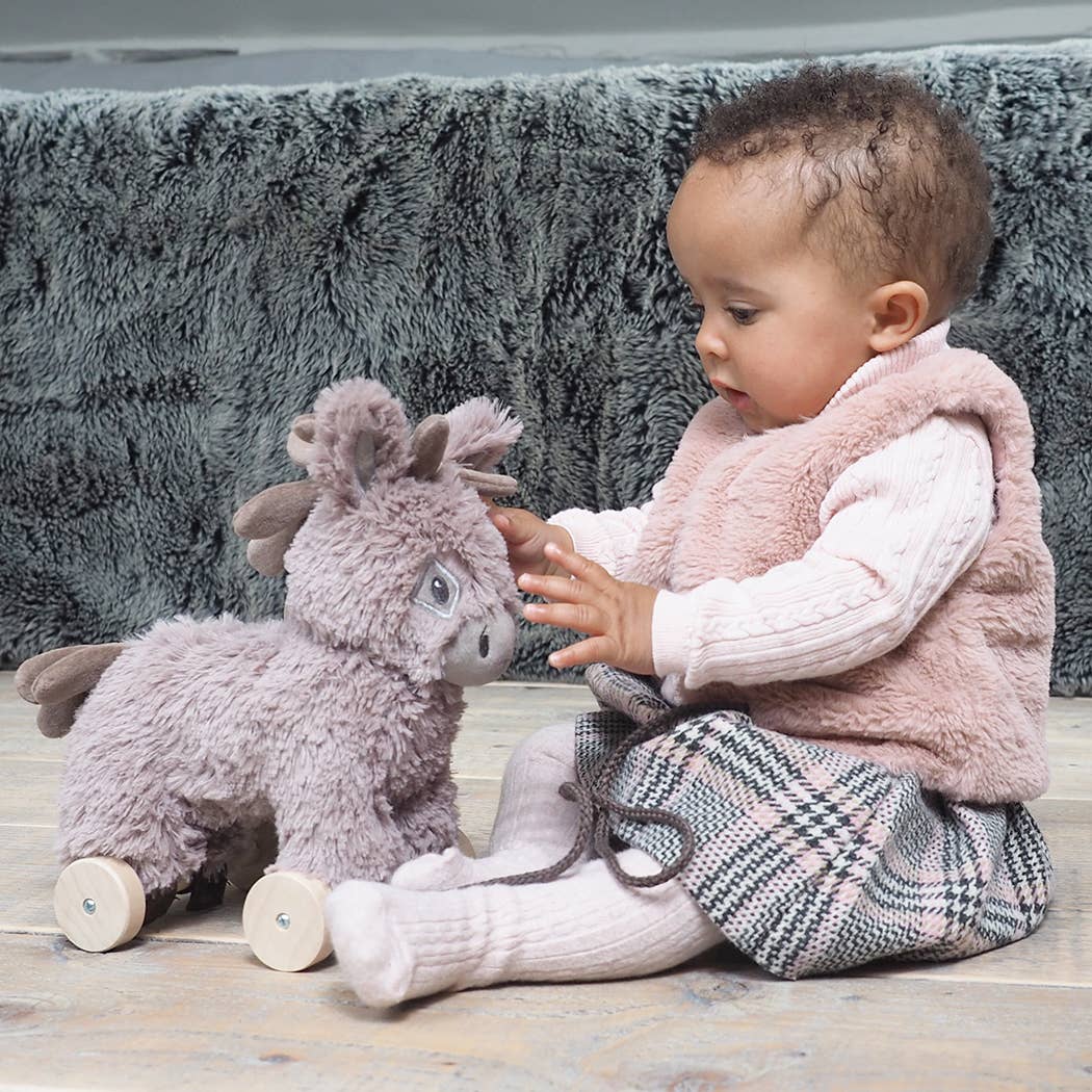 A baby in a pink vest, plaid skirt, and tights sits on a wooden floor, reaching for the Little Bird Told Me UK Norbert Pull Along Baby Toy Donkey to foster motor skills. A fuzzy gray blanket rests in the background.