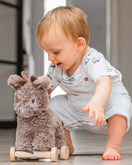 A toddler in light clothes plays on a smooth floor, reaching for the Little Bird Told Me UK Norbert Pull Along Baby Toy Donkey, a fluffy activity toy on wheels that supports motor skills development.