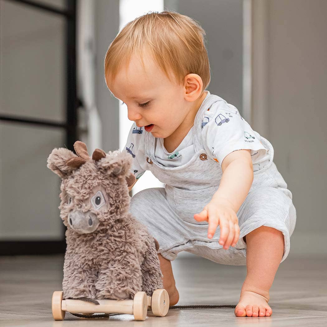 A toddler in light clothes plays on a smooth floor, reaching for the Little Bird Told Me UK Norbert Pull Along Baby Toy Donkey, a fluffy activity toy on wheels that supports motor skills development.