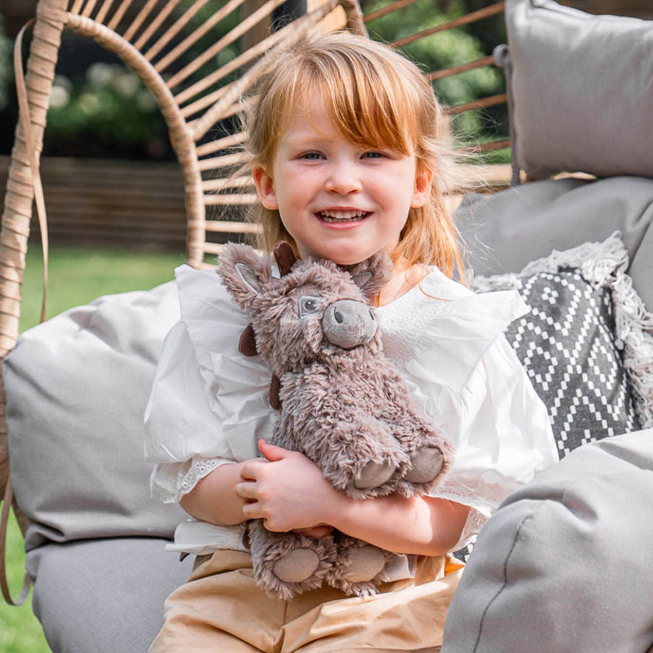 A smiling young girl with red hair, in a white shirt and beige pants, hugs the Pull Along Baby Toy Donkey Norbert by Little Bird Told Me UK while sitting on a cushioned outdoor chair. Greenery and garden furniture are in the background.