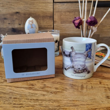 The Cat Mug Alisons Animals Re-charging Cartoon by Alisons Animals, featuring a cat on its back with Im recharging, sits next to its gift box on a wooden surface, surrounded by dried roses and cat-themed decor.