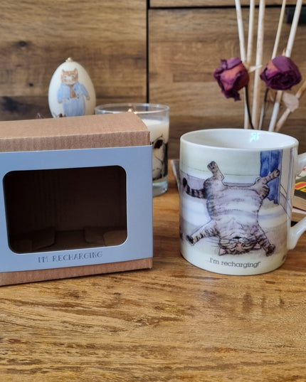 The Cat Mug Alisons Animals Re-charging Cartoon by Alisons Animals, featuring a cat on its back with Im recharging, sits next to its gift box on a wooden surface, surrounded by dried roses and cat-themed decor.