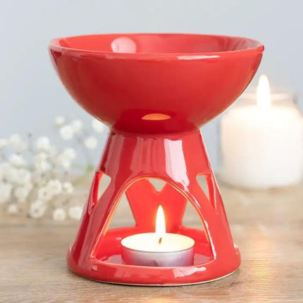 The Something Different Wholesale Red Scented Wax Warmer Oil Burner with a modern geometric design and free melt bar rests on a wooden surface; a tealight candle glows beneath, with blurred white flowers and a candle in the background.