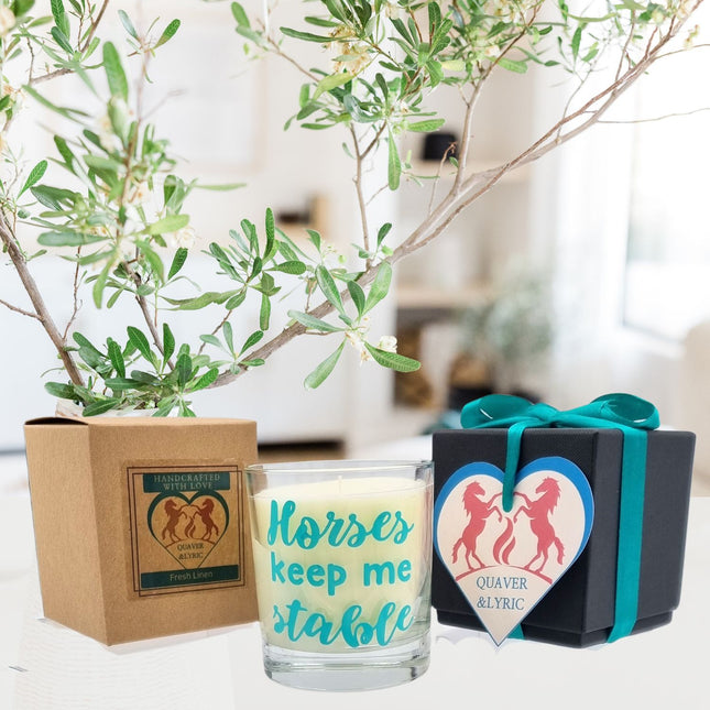 The Quaver&Lyric Scented Candle In Glass Container with Horses Keep Me Stable design sits by brown and blue gift boxes, one with a heart tag featuring two horse silhouettesâ€”an ideal gift for horse lovers. Green leafy branches hang overhead.