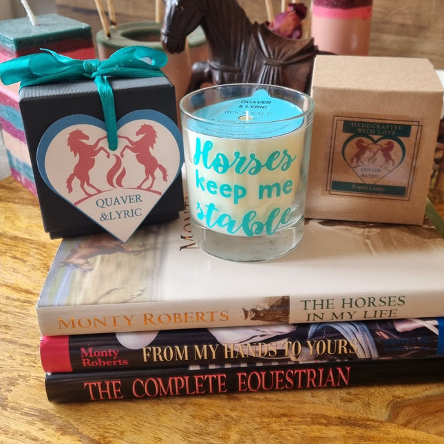 The Quaver&Lyric Scented Candle In Glass Container, featuring the Horses Keep Me Stable design, sits atop equestrian books beside a black gift box with a horse motifâ€”an ideal premium fragrance gift for horse lovers. Carved horses and candles appear in the background.