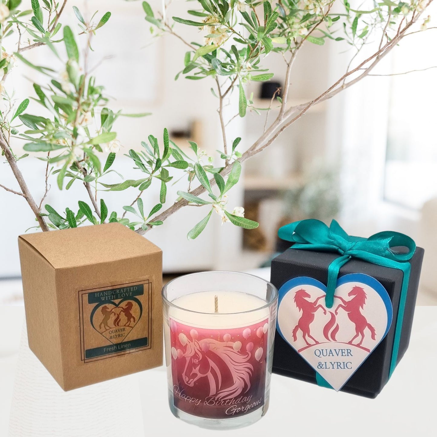 A Scented Candle In Glass Container featuring a Happy Birthday Gorgeous horse design sits by its brown box. Next to it, a Quaver&Lyric gift box with a teal ribbon and heart tag brightens the scene, complemented by a leafy plant.