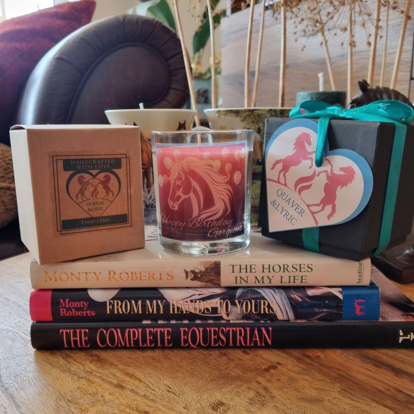 A cozy scene features a stack of equestrian books topped with Quaver&Lyrics Happy Birthday Gorgeous horse design scented candle in glass, plus a Fresh Linen premium fragrance box, couch, cushions, and dried flowers.