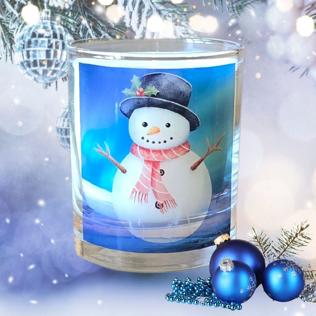 The Quaver&Lyric Christmas Scented Candle features a glass container with a snowman wearing a black hat and red scarf, set amid silver decorations, blue ornaments, pine branches, and festive holiday home decor.